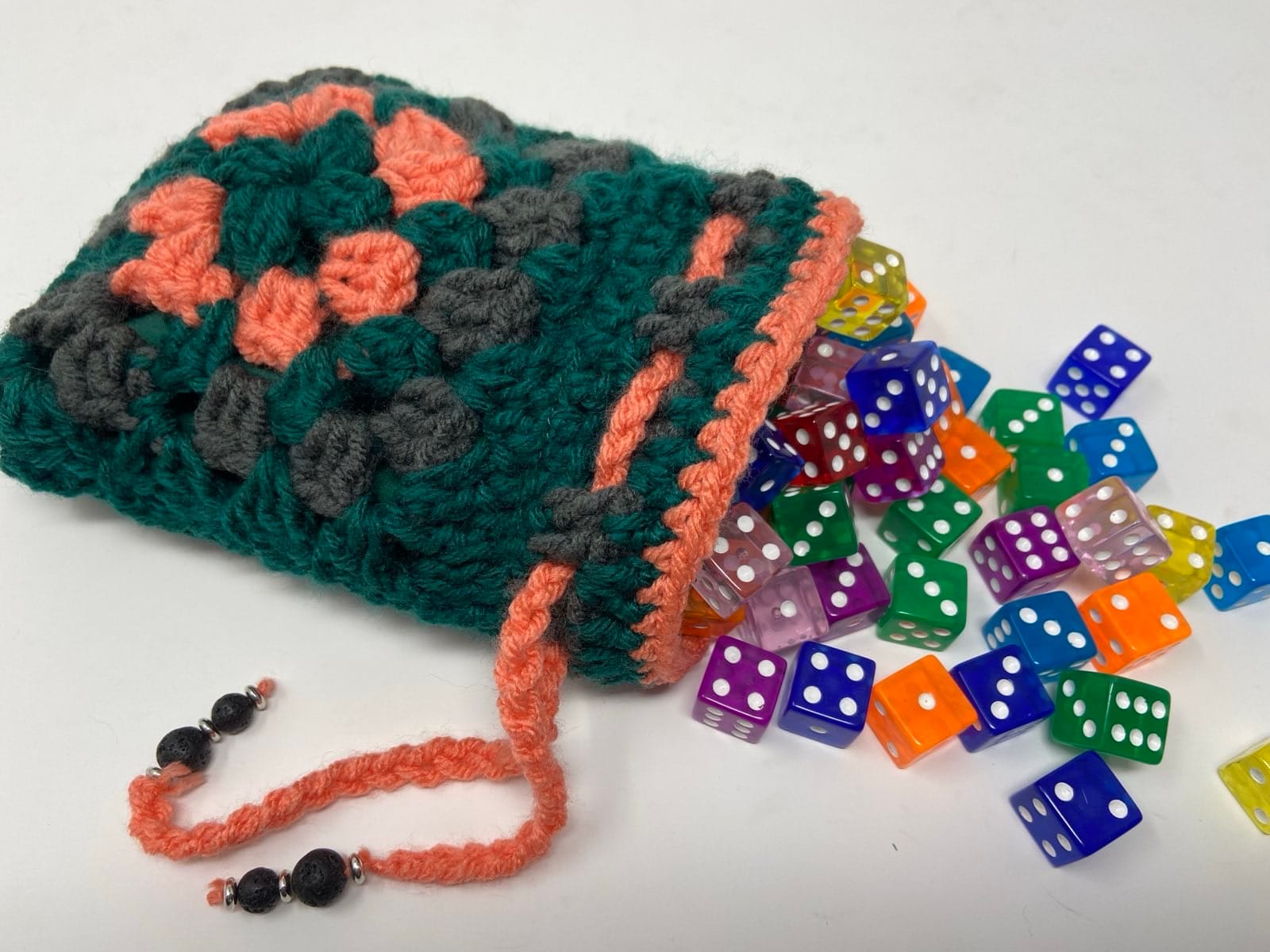 Teal, coral, and gray crocheted dice bag with granny square design, with colorful dice spilling out.