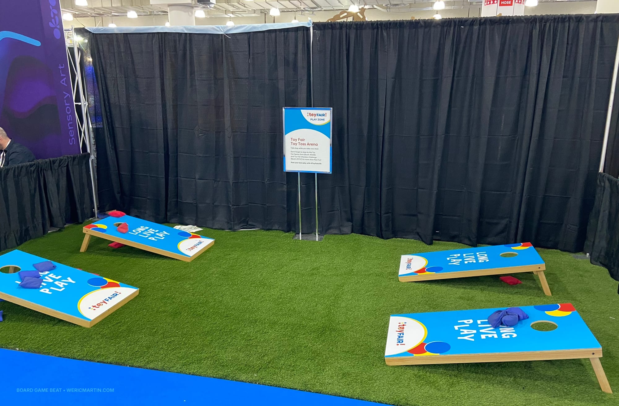Four cornhole boards are in a convention booth that's had artificial turf unrolled across its concrete floor