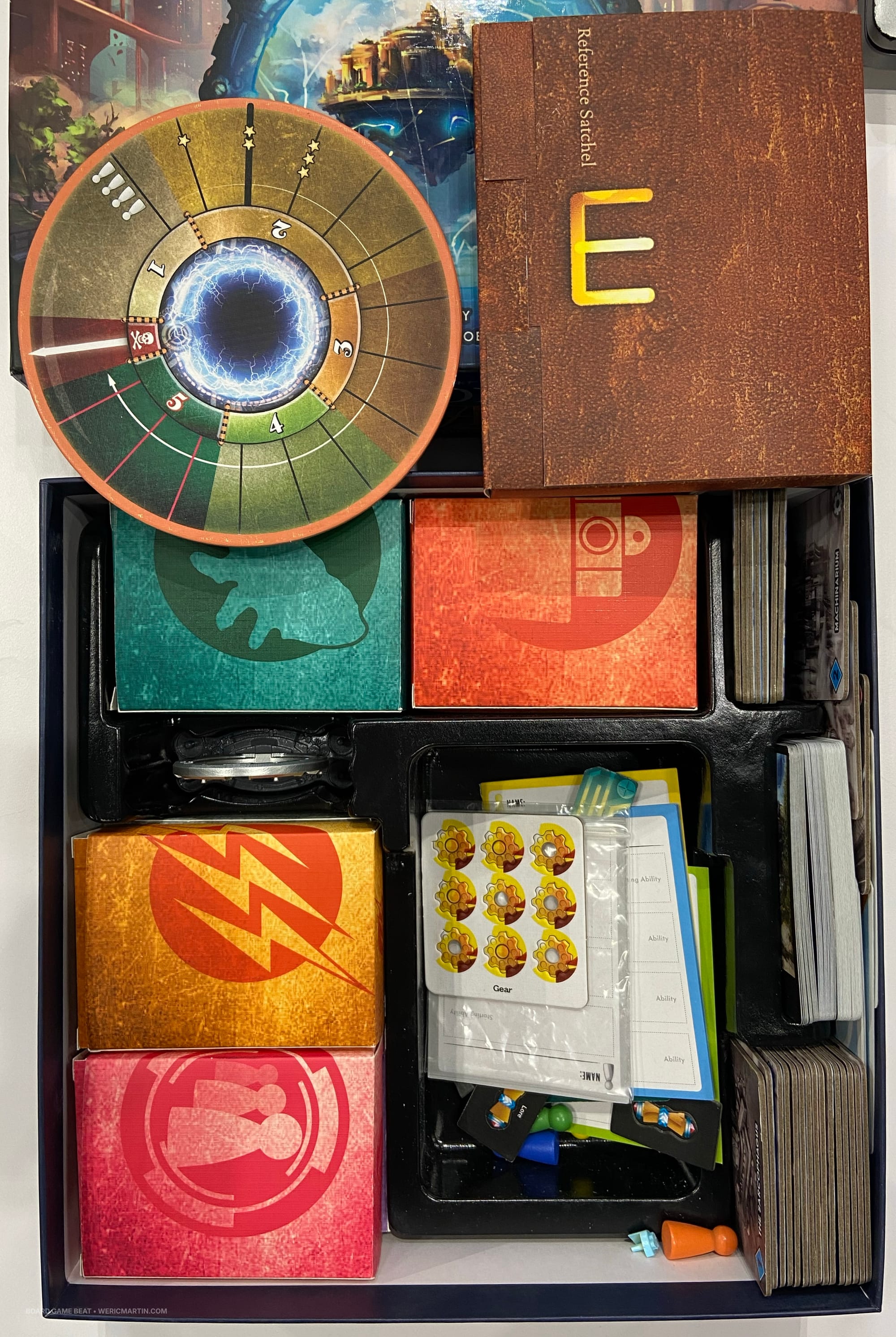 A game tray inside a box holds several closed boxes, with many game components in a well and game tiles and cards standing vertically on the box edge