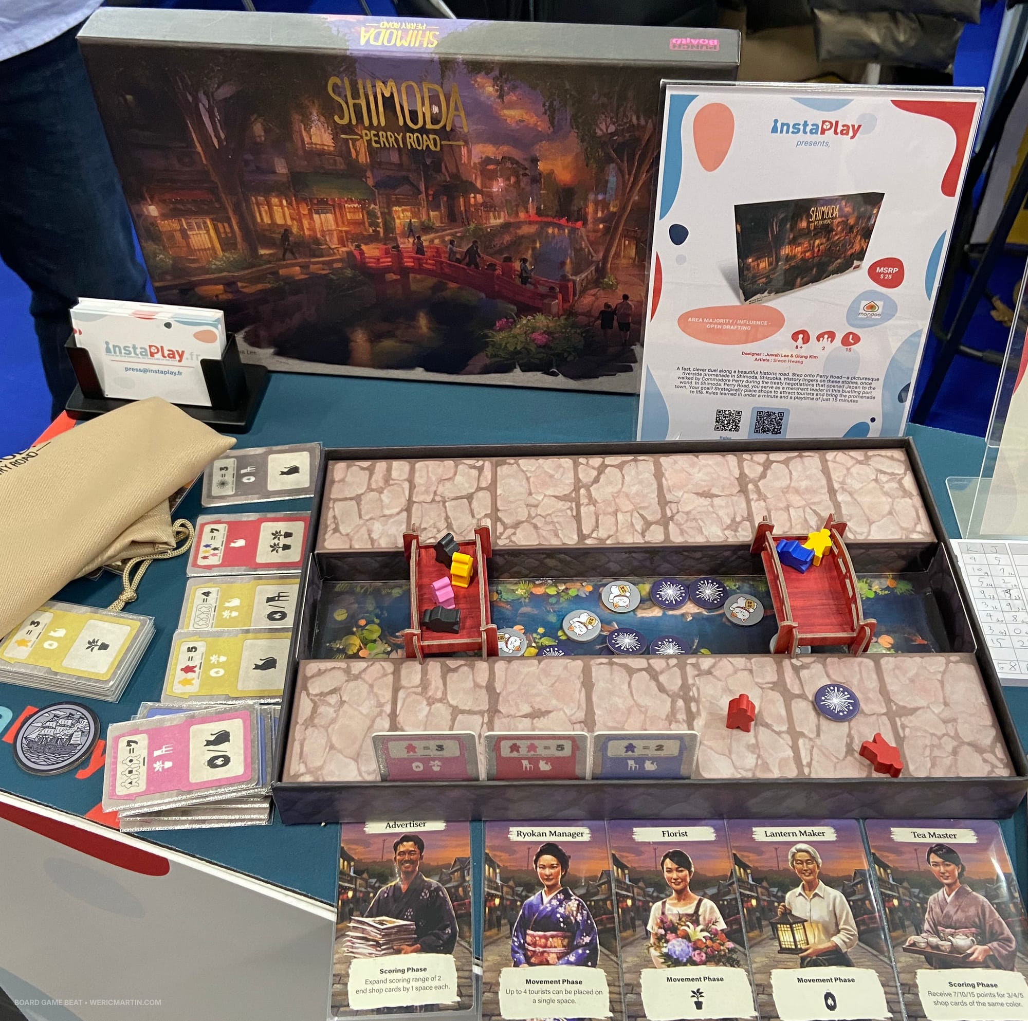 The front cover of Shimota: Perry Road behind the game box, which features two roads that hold tourists; shop cards are placed in the edge of the box to represent buildings