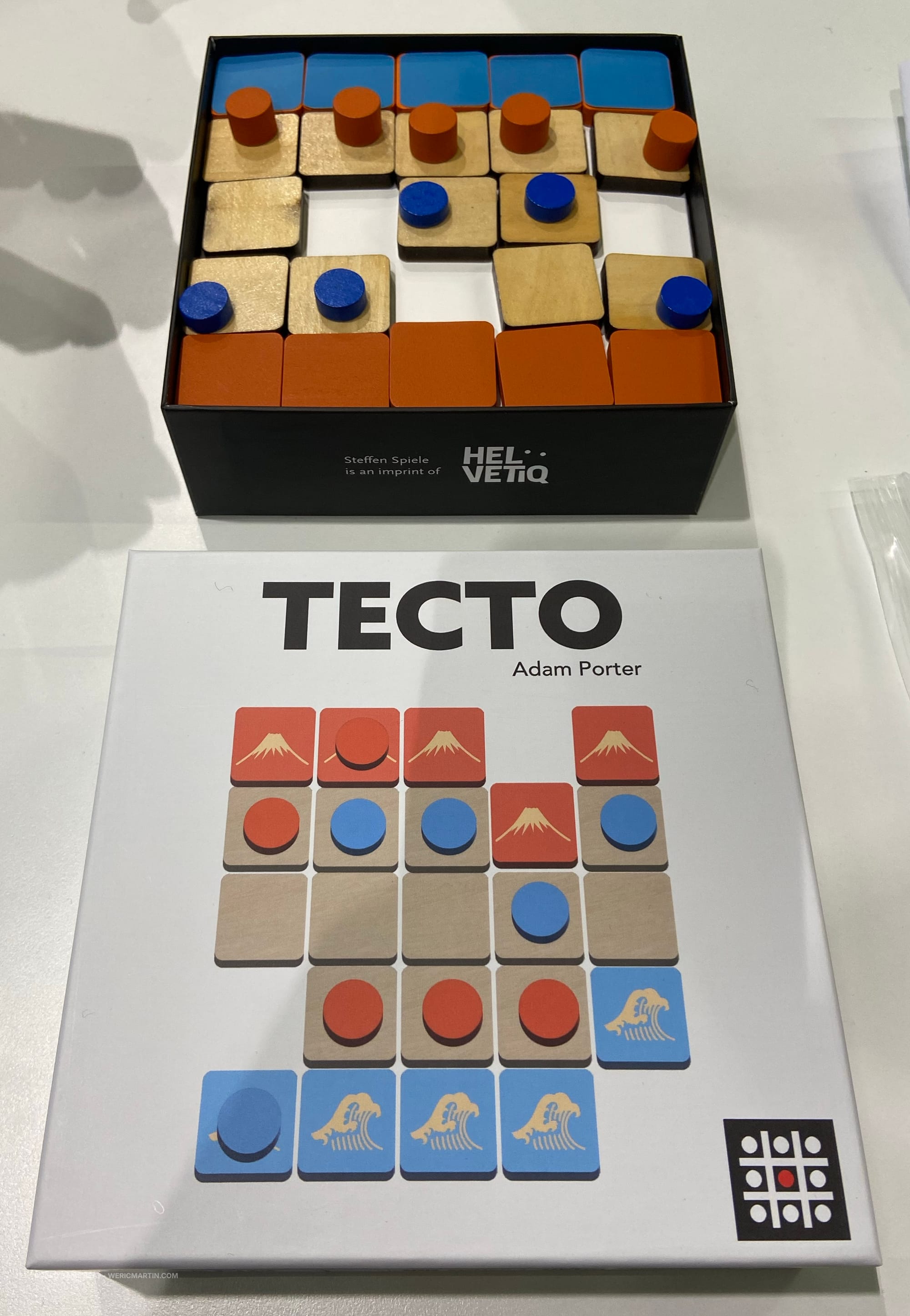 Front cover of Tecto and the game board, which has five blue and five orange discs on tiles of three colors in a 5x5 grid