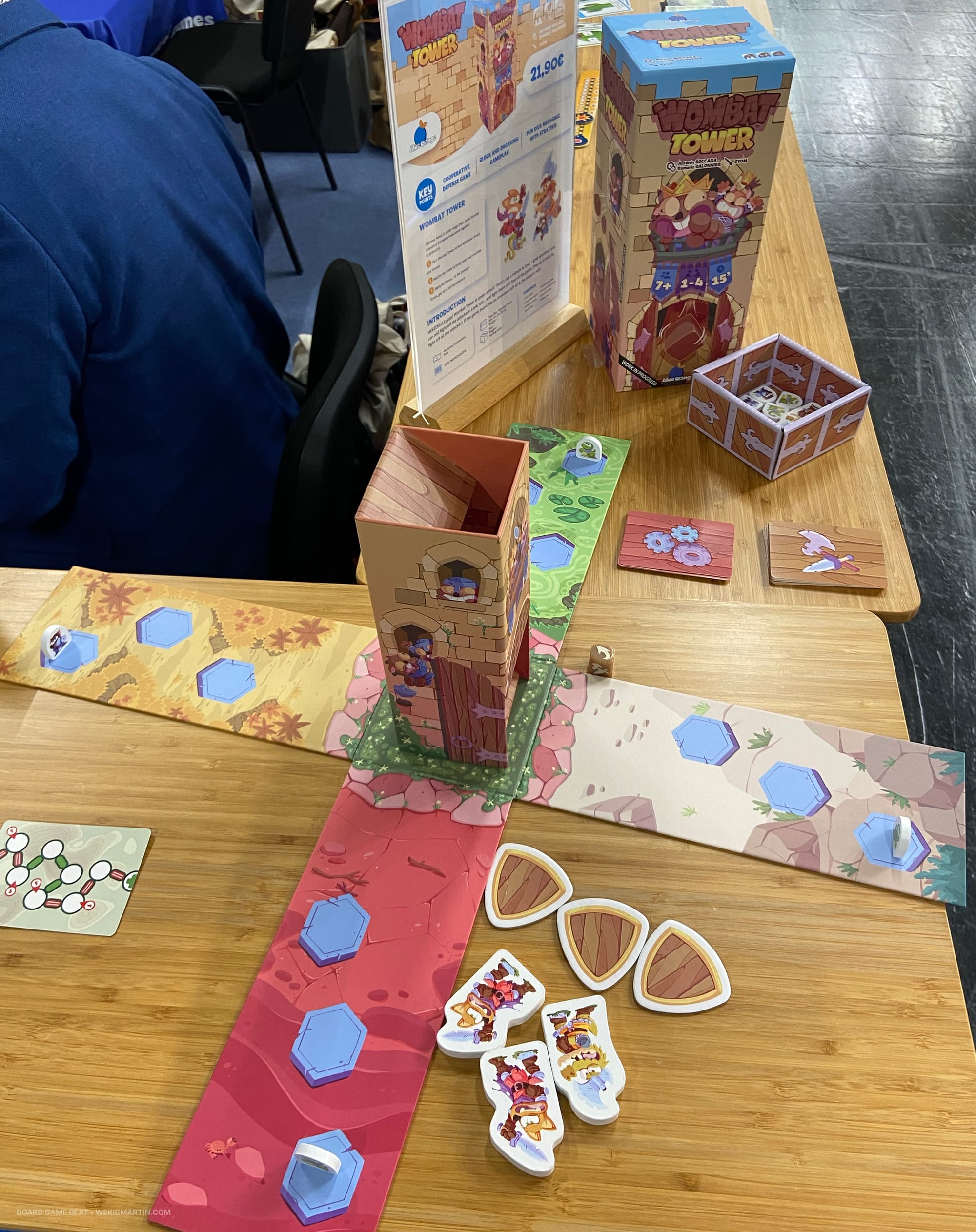 A cardboard dice tower stands in the middle of a cardboard X, with three shields, cards, and tokens nearby