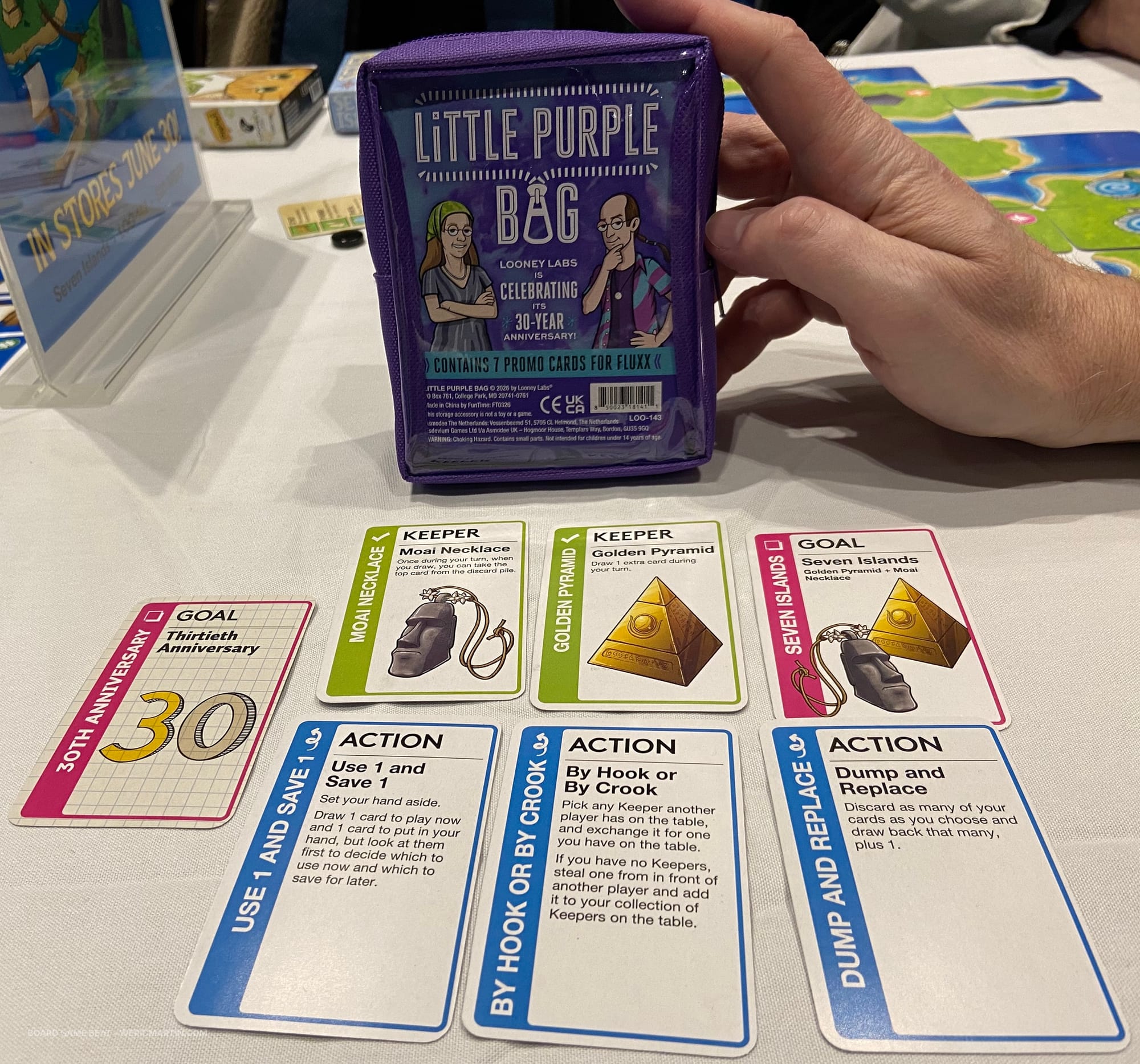 A hand props up a purple Fluxx-sized carrying case behind seven Fluxx promo cards included in this pack