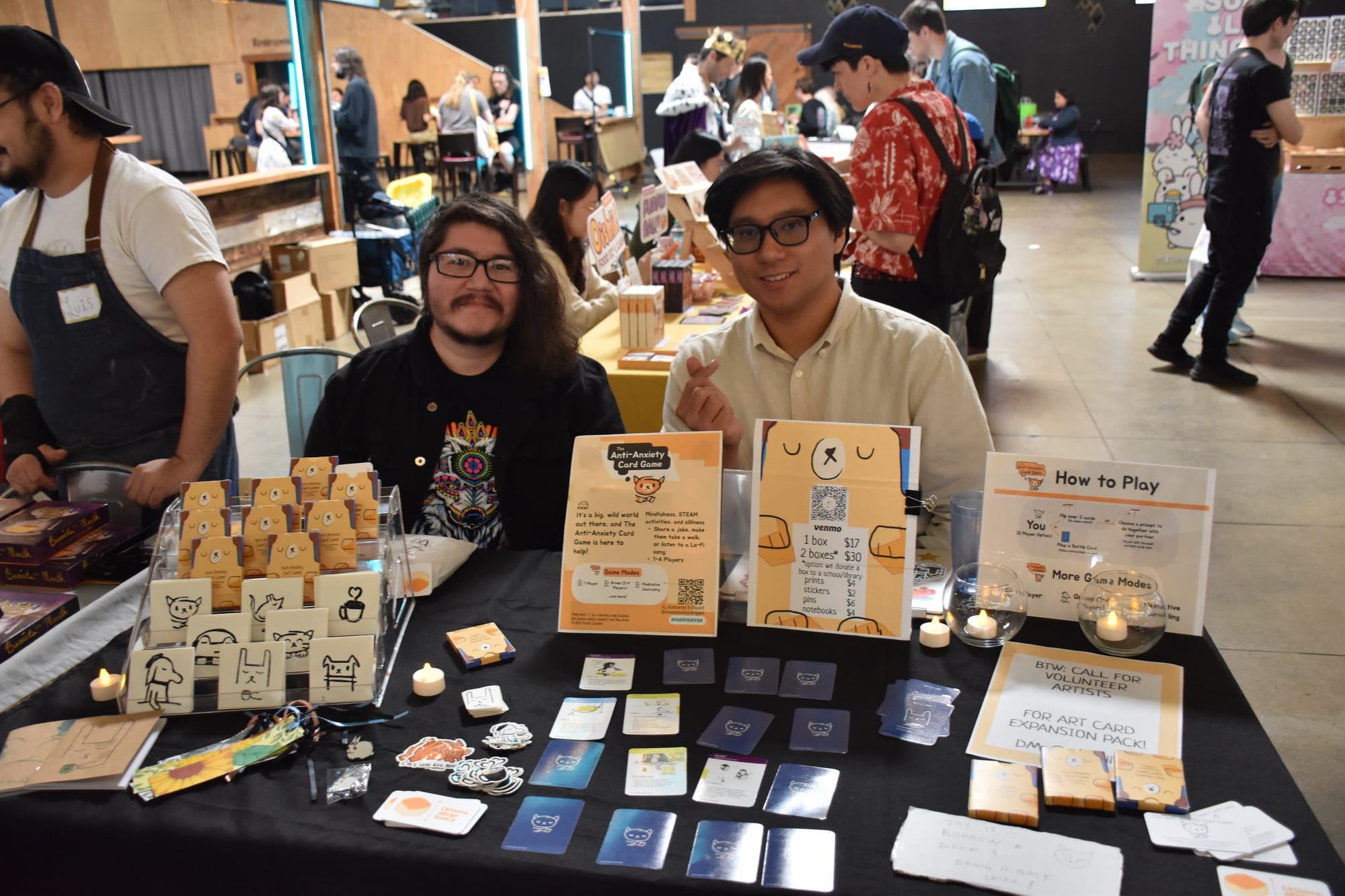 Two people sit behind a table bearing copies of The Anti-Anxiety Card Game, with one of the people making a heart with their fingers; cards, stickers, and pins are spread out on the table, with copies of the game in a plastic rack