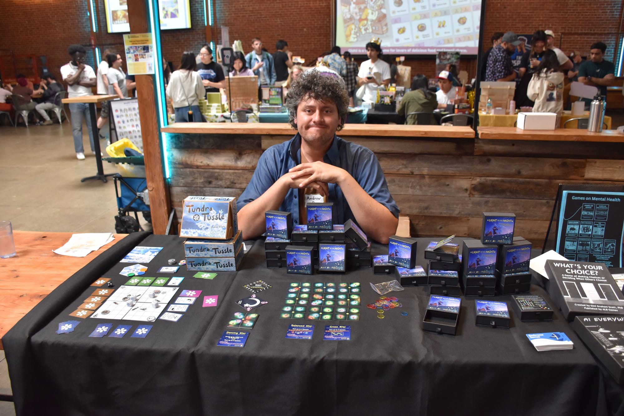 Designer Andrew DiLullo sits behind a table bearing copies of Light Up the Night, with tiles arranged in a 6x6 grid and other components surrounding those tiles; another game, Tundra Tussle, is adjacent to this game