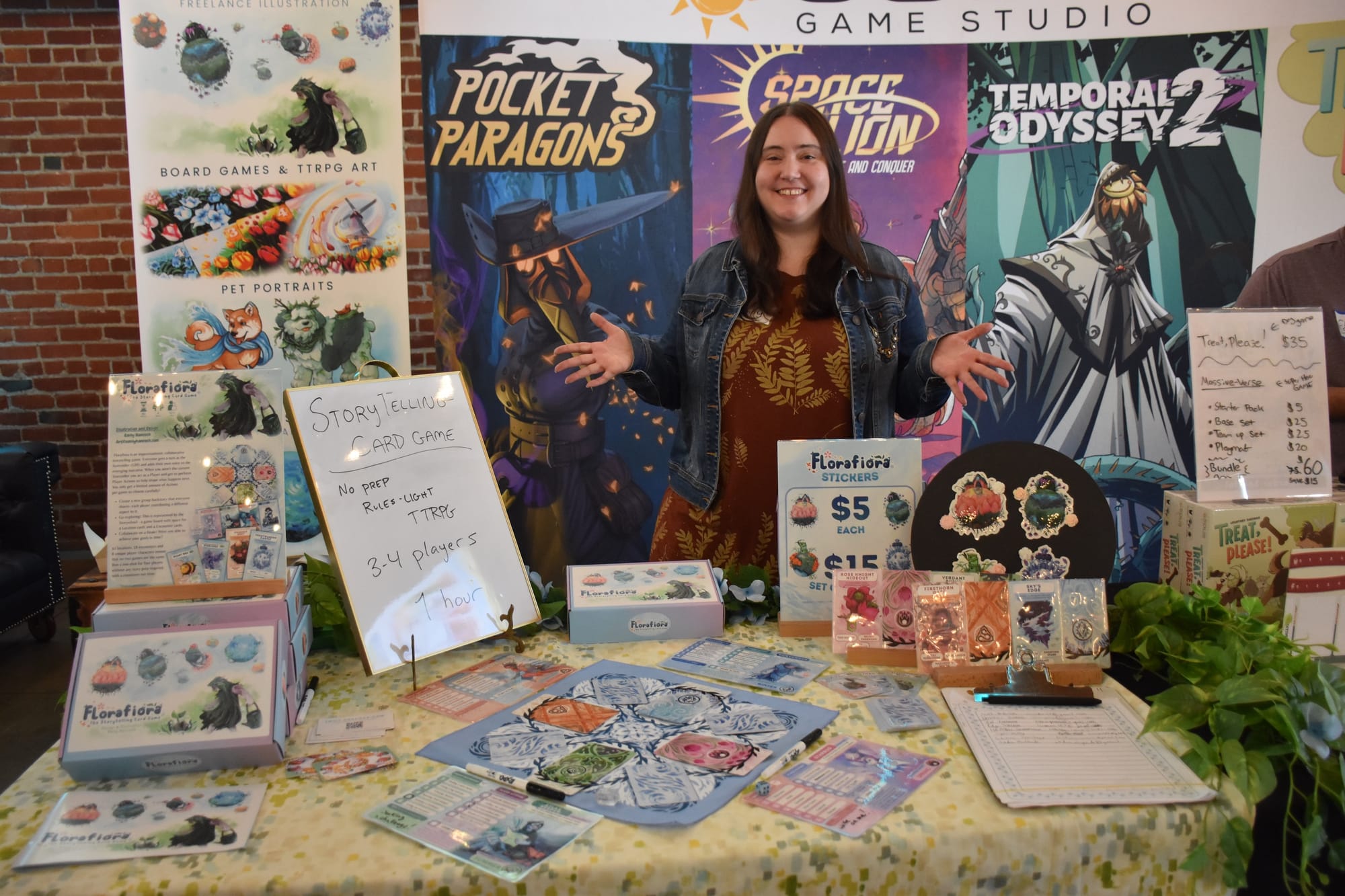 Designer/artist Emily Hancock stands behind a table bearing her game Florafiora, which has a central game board with cards on it and a player board on each side; stickers and copies of the game are available