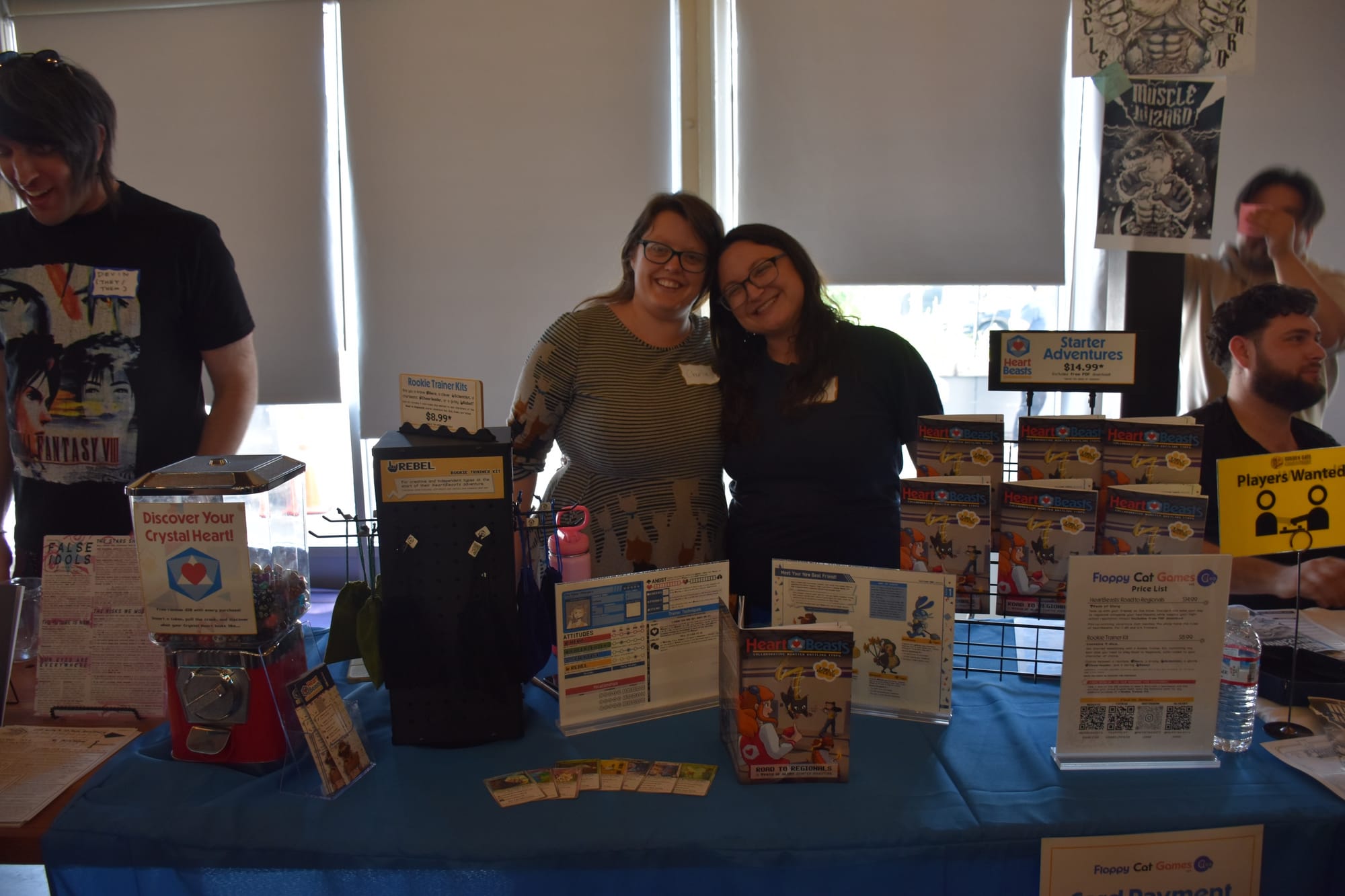 Two people stand behind a table bearing copies of the RPG HeartBeasts, as well as promotional signs and dice available for purchase